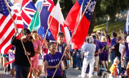 A group of laughing students carry flags of different nations in a TCU Homecoming parade, led by a girl wearing a shirt that says "Frogs"