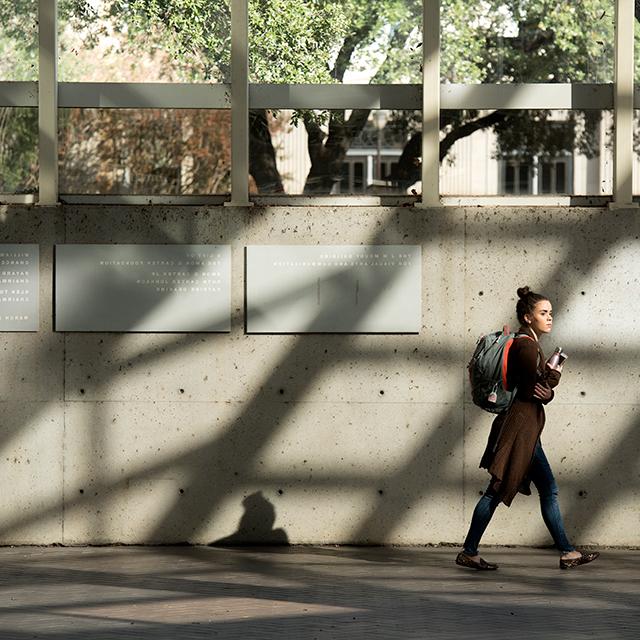 A student carrying a backpack walks through the Moudy Building as the structure's architectural features cast shadows dramatically on a concrete wall
