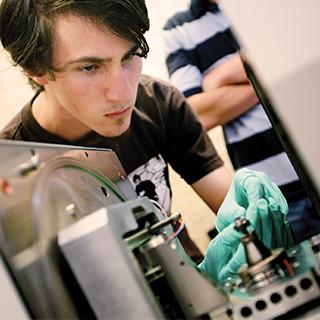 A male student wearing green protective gloves works on a piece of electronics